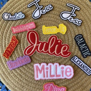 May include: A collection of embroidered patches with various names and phrases. The patches feature names like "Jess", "Julie", and "Millie", along with phrases such as "Lemon-Up" and "Galactic". The patches are in various colors, including red, pink, and yellow.