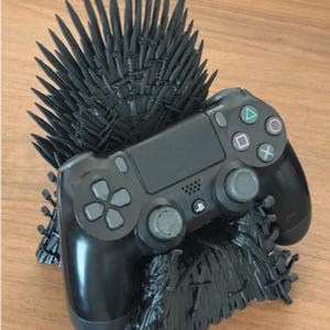 Op de afbeelding: Een zwarte plastic gaming controller standaard ontworpen om te lijken op een troon gemaakt van zwaarden. De standaard heeft een hoge, stekelige rugleuning en een basis opgebouwd uit talloze zwaardgrepen en bladen, die een zwarte draadloze controller vasthoudt.
