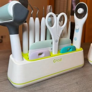 Könnte beinhalten: Ein weiß-grüner Cricut-Werkzeugorganizer mit verschiedenen Bastelwerkzeugen. Der Organizer enthält eine Rolle, eine Schere, Schaber und andere Werkzeuge. Das Wort "Cricut" ist auf der Vorderseite aufgedruckt.