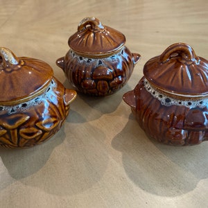 May include: Three brown ceramic pots with lids. Each pot has a decorative design of mushrooms and a white lace trim around the rim.