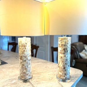 May include: Two matching table lamps with beige fabric shades. The cylindrical bases are covered in a mosaic of small, rectangular pieces in shades of cream, beige, and brown. The lamps sit on clear, circular bases and have silver-toned hardware.