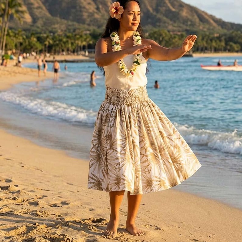 Op de afbeelding: Een vrouw in een witte top en een rok met een beige en wit palmbladpatroon, die een traditionele dans uitvoert op een zandstrand. Ze draagt een bloem in haar haar en een lei.