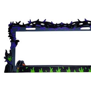 Op de afbeelding: Een kentekenplaathouder met een donkergrijze basis en zwarte en paarse accenten. Het frame is versierd met een draak, een heks en een doornig wijnstokontwerp. Groene vlammen bevinden zich aan de onderkant van het frame.