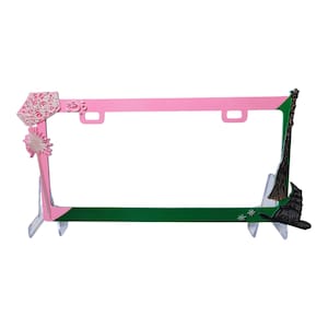 May include: A pink and green license plate frame with decorative accents. The frame has a pink top and a green bottom. Embellishments include a witch's hat, a broom, and floral designs. The frame is designed to hold a standard license plate.