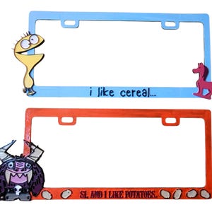 May include: Two license plate frames. The top frame is light blue with the text "i like cereal..." and a cartoon character and a pink horse. The bottom frame is orange with the text "SL AND I LIKE POTATOES" and a cartoon monster.