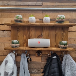 May include: A rustic wooden wall shelf with two tiers, featuring four metal hooks and a bottle opener. The top shelf holds two glass terrariums and two white heart-patterned pots. The bottom shelf displays two glass terrariums and a white JBL speaker.