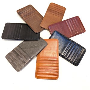 May include: A collection of seven leather cardholders in various colors, including black, brown, and gray. Each cardholder has multiple slots for cards and a rectangular shape. The cardholders are arranged in a circular pattern against a white background.