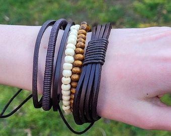 May include: A set of five brown leather bracelets with a beaded bracelet in the center. The beaded bracelet has brown and white beads.