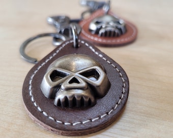 May include: A brown leather keychain with a silver skull charm. The keychain has white stitching around the edges.