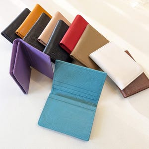 May include: A collection of leather cardholders in various colors, including black, yellow, tan, red, brown, white, and blue. One purple cardholder is open, displaying the interior. The cardholders are arranged on a white surface.