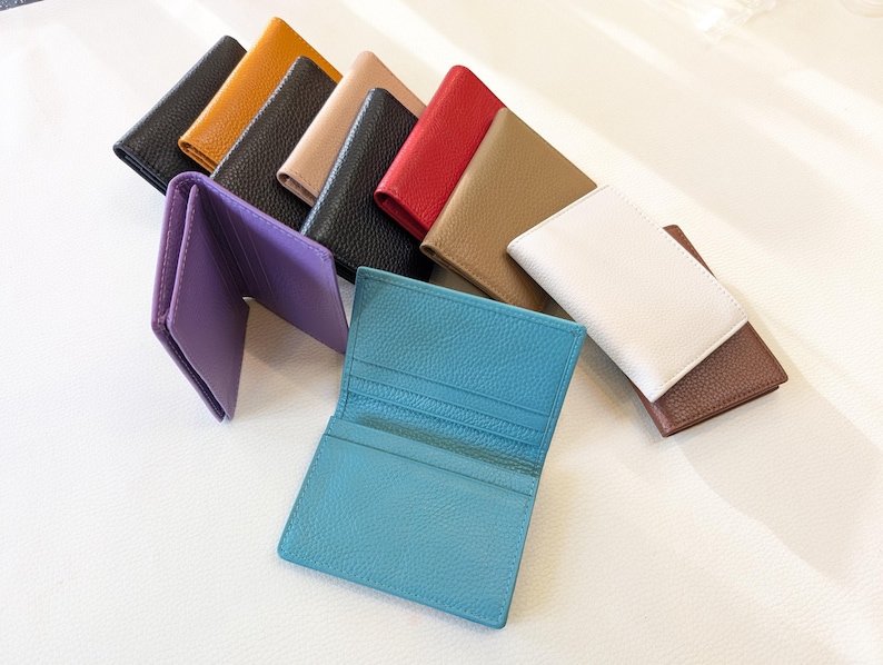 May include: A collection of leather cardholders in various colors, including black, yellow, tan, red, brown, white, and blue. One purple cardholder is open, displaying the interior. The cardholders are arranged on a white surface.