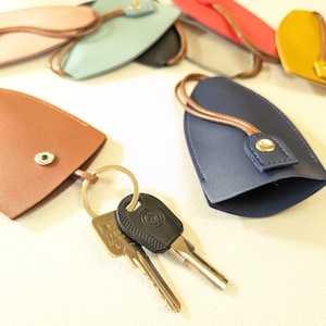 May include: A set of keychains made of leather in various colors, including brown, blue, pink, and yellow. The keychains have a loop for attaching keys and a small button closure. Two keys are attached to one of the keychains.
