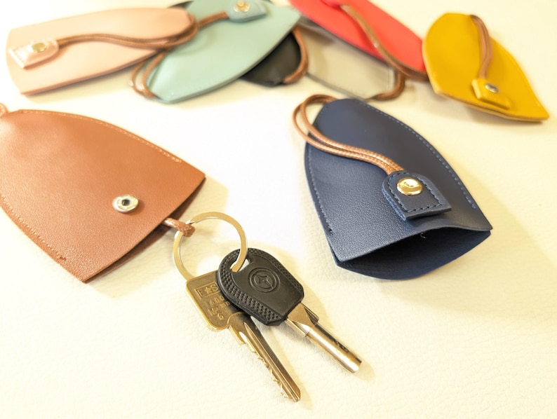 May include: A set of keychains made of leather in various colors, including brown, blue, pink, and yellow. The keychains have a loop for attaching keys and a small button closure. Two keys are attached to one of the keychains.