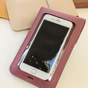 May include: A burgundy leather phone case with a clear plastic window to display a smartphone. The case has a stitched edge and a small hole at the bottom.
