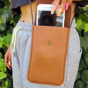 May include: A brown leather phone case with a strap. The case has a flap that closes with a snap. A smartphone is visible inside the case.