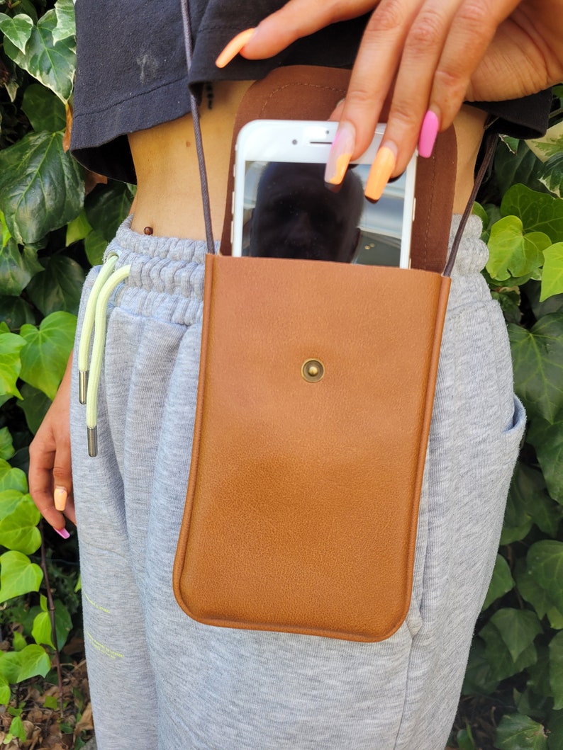 May include: A brown leather phone case with a strap. The case has a flap that closes with a snap. A smartphone is visible inside the case.