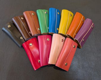 May include: A collection of 14 leather keyrings in various colours, including black, brown, red, pink, green, blue, yellow, and purple. The keyrings are arranged in a semi-circle, with the tips of the keyrings pointing towards the centre.