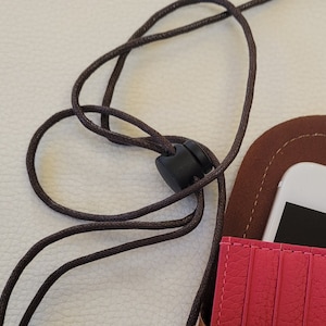 May include: A pink leather phone case with a brown leather flap and a black adjustable strap. The case has a slot for a phone and a card holder.