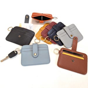 May include: A collection of leather cardholders in various colors, including black, brown, blue, gray, yellow, and burgundy. Each cardholder has a key ring and a snap closure. Some have a small strap.