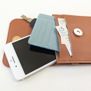 May include: A brown leather wallet with a snap closure, containing a blue leather card holder, a smartphone, keys, and British currency. The wallet has a pocket for cash and a separate compartment for cards.