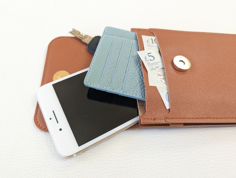 May include: A brown leather wallet with a snap closure, containing a blue leather card holder, a smartphone, keys, and British currency. The wallet has a pocket for cash and a separate compartment for cards.