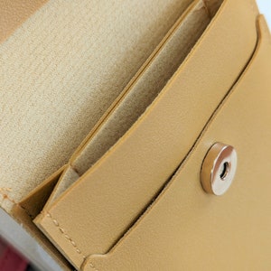 May include: A tan leather wallet with a flap closure and a silver snap button. The wallet has a small pocket on the outside and a larger pocket on the inside.