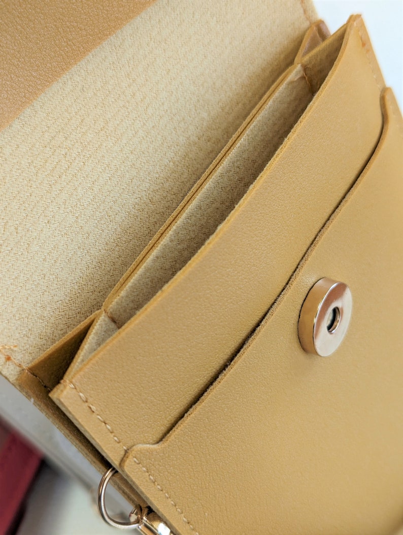 May include: A tan leather wallet with a flap closure and a silver snap button. The wallet has a small pocket on the outside and a larger pocket on the inside.