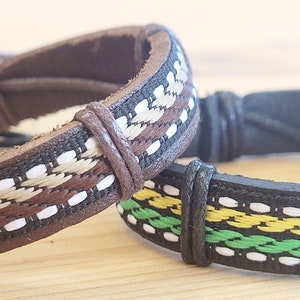 Könnte beinhalten: Zwei Lederarmbänder mit gewebten Mustern. Ein Armband ist braun mit gewebten Streifen in Tan und Weiß. Das andere Armband ist schwarz mit gewebten Streifen in Grün, Gelb und Weiß.