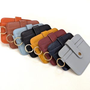 May include: A collection of colorful leather cardholders with key rings. The cardholders are in various colors, including orange, brown, blue, black, yellow, burgundy, navy, and gray. Each cardholder has a gold-colored key ring attached.