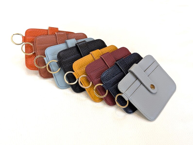 May include: A collection of colorful leather cardholders with key rings. The cardholders are in various colors, including orange, brown, blue, black, yellow, burgundy, navy, and gray. Each cardholder has a gold-colored key ring attached.
