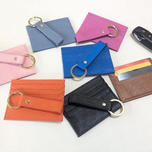 May include: Assortment of nine leather card holders with key rings. The card holders are in various colors, including pink, blue, brown, black, and orange. Each card holder has a gold key ring attached.