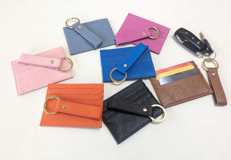 May include: Assortment of nine leather card holders with key rings. The card holders are in various colors, including pink, blue, brown, black, and orange. Each card holder has a gold key ring attached.