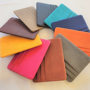 May include: A set of ten leather card holders in various colors, including brown, gray, orange, yellow, pink, blue, teal, and black. The card holders are arranged in a circle on a white surface.