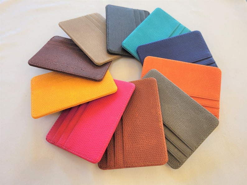 May include: A set of ten leather card holders in various colors, including brown, gray, orange, yellow, pink, blue, teal, and black. The card holders are arranged in a circle on a white surface.