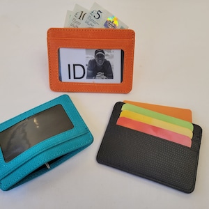May include: Three different card holders, one orange, one teal, and one black. The orange card holder has a clear window for an ID card and is filled with cash. The teal card holder has a clear window for an ID card. The black card holder has four slots for cards.