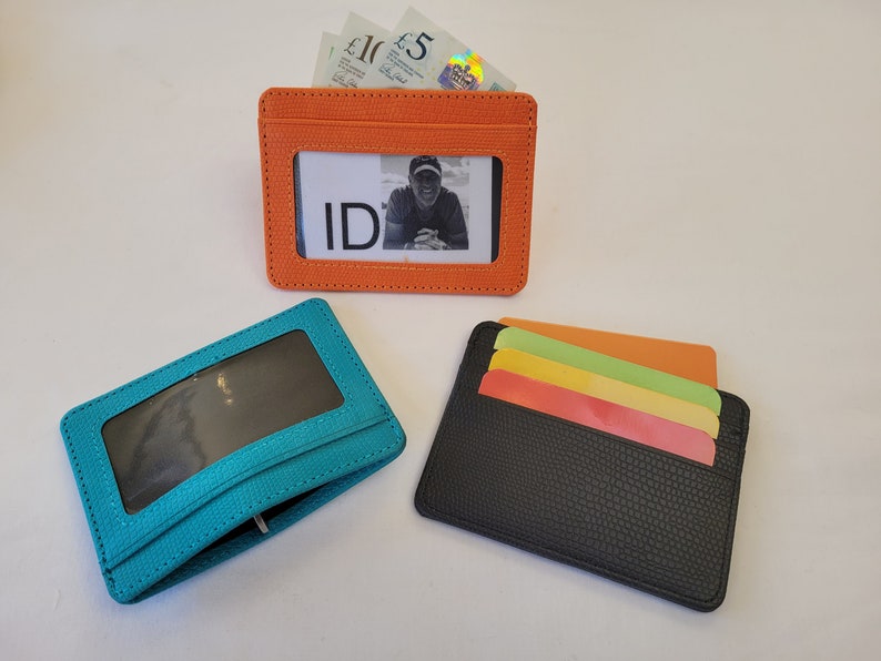 May include: Three different card holders, one orange, one teal, and one black. The orange card holder has a clear window for an ID card and is filled with cash. The teal card holder has a clear window for an ID card. The black card holder has four slots for cards.