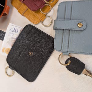 May include: A collection of leather cardholders in various colors, including black, blue, and yellow. Each cardholder features a key ring and a snap closure. One cardholder holds a twenty-pound note.