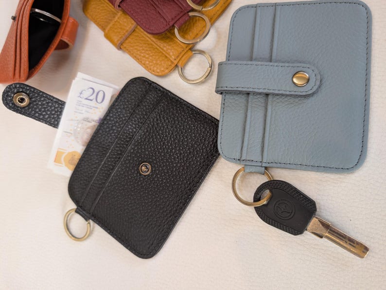 May include: A collection of leather cardholders in various colors, including black, blue, and yellow. Each cardholder features a key ring and a snap closure. One cardholder holds a twenty-pound note.