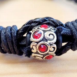 Puede incluir: Un brazalete de cuero negro con una cuenta de plata con diamantes de imitación rojos. La cuenta tiene un diseño decorativo de espiral.