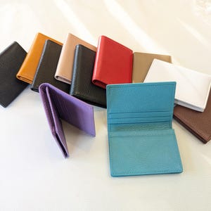 May include: A collection of leather cardholders in various colors, including black, tan, red, and white. One purple cardholder is open, displaying the interior. A turquoise cardholder is also open, showcasing its compartments. The cardholders are arranged on a white surface.