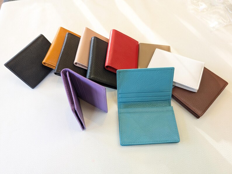 May include: A collection of leather cardholders in various colors, including black, tan, red, and white. One purple cardholder is open, displaying the interior. A turquoise cardholder is also open, showcasing its compartments. The cardholders are arranged on a white surface.