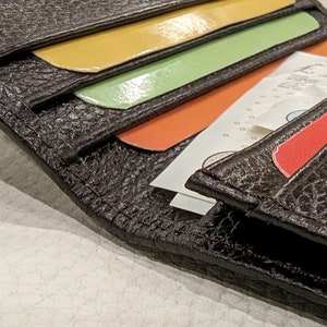 May include: Close-up of a dark brown leather wallet with a textured surface. The wallet is open, revealing colorful cards in yellow, green, and orange, along with folded paper money and a red card. The wallet is a practical accessory.