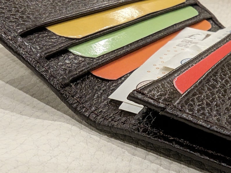 May include: Close-up of a dark brown leather wallet with a textured surface. The wallet is open, revealing colorful cards in yellow, green, and orange, along with folded paper money and a red card. The wallet is a practical accessory.