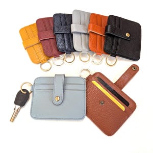 May include: A collection of leather cardholders in various colors, including yellow, burgundy, navy, gray, orange, and black. Each cardholder has a key ring and a snap closure. One light blue cardholder has a key attached, and a brown cardholder is open, displaying cards.