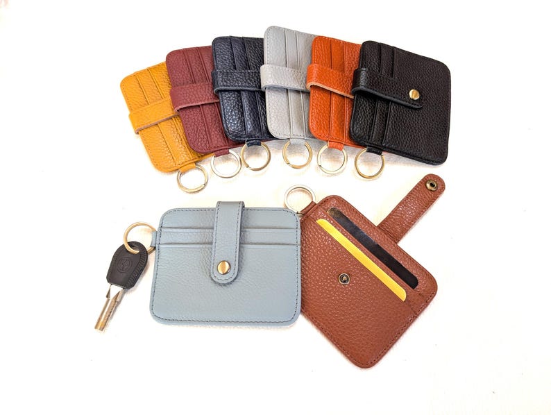 May include: A collection of leather cardholders in various colors, including yellow, burgundy, navy, gray, orange, and black. Each cardholder has a key ring and a snap closure. One light blue cardholder has a key attached, and a brown cardholder is open, displaying cards.