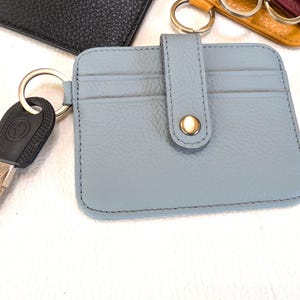 May include: A light blue leather cardholder with a snap closure and key ring. The cardholder has two card slots and is attached to a silver key ring with a car key. Other colored cardholders are visible in the background.