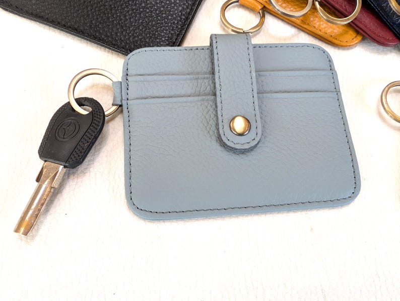 May include: A light blue leather cardholder with a snap closure and key ring. The cardholder has two card slots and is attached to a silver key ring with a car key. Other colored cardholders are visible in the background.