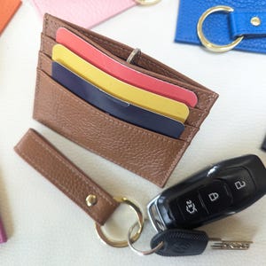 May include: A brown leather card holder with a key ring attached. The card holder has three card slots and is made of a textured leather. The key ring has a silver key ring and a silver key chain.