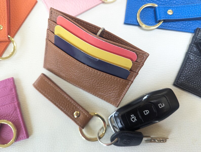 May include: A brown leather card holder with a key ring attached. The card holder has three card slots and is made of a textured leather. The key ring has a silver key ring and a silver key chain.