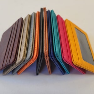 May include: A stack of ten leather card holders in various colors, including brown, orange, gray, blue, teal, and yellow. The card holders are folded in half and arranged in a pyramid shape.